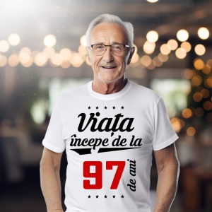 Bărbat în vârstă purtând ochelari și un tricou alb cu mesajul 'Viața începe de la 97 de ani', într-un cadru iluminat cald.