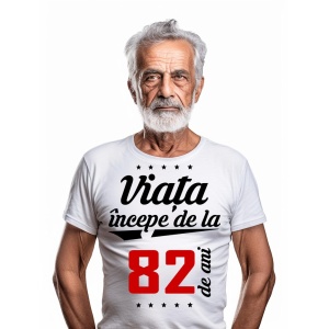 Bărbat în vârstă cu barbă albă, purtând un tricou alb cu mesajul 'Viața începe de la 82 de ani', pe un fundal alb simplu.