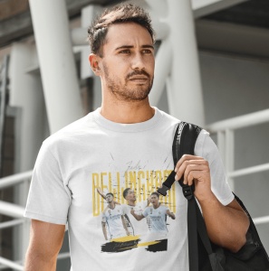 Un bărbat poartă un tricou alb cu un design grafic dedicat jucătorului de fotbal Jude Bellingham, incluzând imagini ale acestuia în acțiune și numele său scris cu litere galbene mari. Bărbatul ține o geantă pe umăr.
