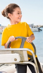 Fată zâmbitoare purtând un tricou galben, conducând o barcă într-o zi însorită, cu apă și bărci ancorate în fundal