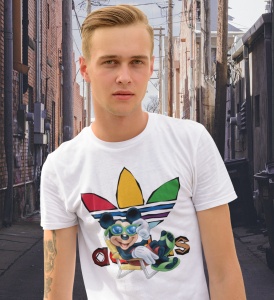 Tânăr cu păr blond și privire serioasă, purtând un tricou alb cu un design grafic colorat ce include un personaj animat și elemente stilizate, aflat pe o stradă urbană îngustă, înconjurată de clădiri de cărămidă și fire electrice.