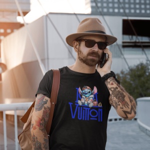 Bărbat purtând o pălărie și ochelari de soare, cu un tricou negru pe care este imprimat un personaj animat albastru(Stitch) și un logo stilizat Louis Vuitton, vorbind la telefon și având un rucsac maro pe umăr și tatuaje pe braț.