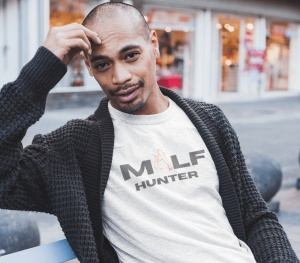 Un bărbat relaxat poartă un tricou alb cu textul 'MILF HUNTER' în font gri și o ilustrație stilizată în roșu, alături de un cardigan gros. Fundalul este un spațiu urban, cu vitrine de magazine în spate.