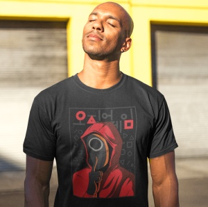 Un bărbat purtând un tricou negru cu un design inspirat din Squid Game, înfățișând un personaj cu mască roșie și simboluri geometrice, pozând în lumina soarelui în fața unui fundal galben.