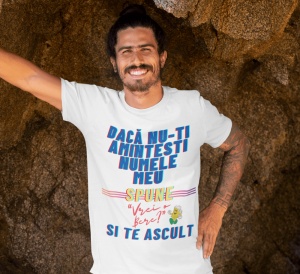 Un bărbat zâmbitor poartă un tricou alb cu mesajul amuzant 'Dacă nu-ți amintești numele meu, spune „Vrei bere?” și te ascult', colorat în albastru, galben și roșu. Bărbatul are părul prins și stă sprijinit pe o suprafață de stâncă.