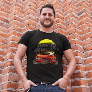 Bărbat zâmbitor purtând un tricou negru cu un design grafic care prezintă o mașină sport roșie și textul 'Why so negative?' în fața unui fundal galben circular, stând în fața unui perete din cărămidă roșie.