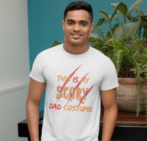 Un bărbat care zâmbește, purtând un tricou alb cu textul 'This is my scary dad costume' în nuanțe de portocaliu și roșu, completat cu un design de zgârieturi de gheare. Bărbatul stă în fața unor plante verzi într-un decor interior.