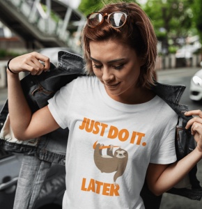 Tânără purtând un tricou alb cu un design care înfățișează un leneș atârnând de o ramură și textul 'JUST DO IT. LATER' scris în portocaliu. Ea poartă o jachetă de blugi descheiată, care este trasă pe umeri, și privește în jos cu un zâmbet subtil. Fundalul