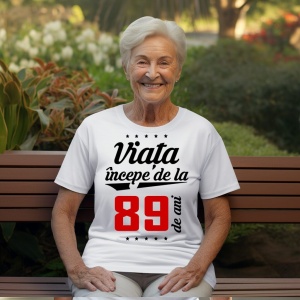 O femeie purtând un tricou alb cu textul  'Viața începe de la 89 de ani'. Fundal din lemn cu diferite nuanțe de maro și verde.
