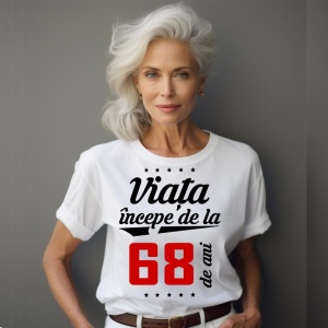 O femeie purtând un tricou alb cu textul  'Viața începe de la 68 de ani'. Fundal din lemn cu diferite nuanțe de maro și verde.