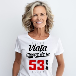 O femeie purtând un tricou alb cu textul  'Viața începe de la 53 de ani'. Fundal din lemn cu diferite nuanțe de maro și verde.