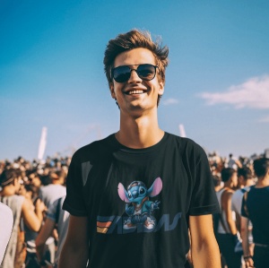 Tânăr zâmbitor la un eveniment în aer liber, purtând ochelari de soare și un tricou negru cu un design colorat care include un personaj animat albastru(Stitch) și logo-ul Adidas stilizat. În fundal se vede o mulțime de oameni sub un cer senin.