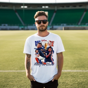 Bărbat purtând un tricou alb cu un design dedicat lui Kylian Mbappé, incluzând imagini ale fotbalistului, textul 'Allez les Bleus!' și steagul Franței, pozând pe un teren de fotbal.