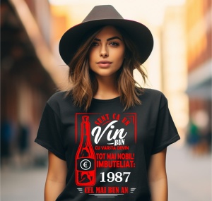 Femeie zâmbitoare purtând un tricou negru cu un mesaj în română despre vin, în care se menționează că este ca un vin bun care devine mai nobil cu vârsta, îmbuteliat în 1987, cel mai bun an.