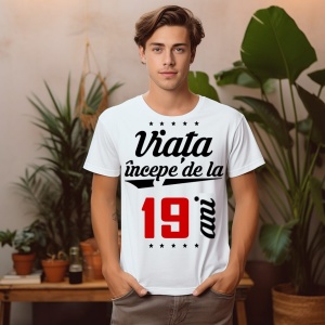 Bărbat tânăr purtând un tricou alb cu mesajul 'Viața începe de la 19 ani', stând într-un mediu decorat cu plante.