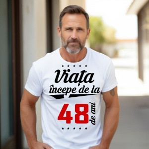 Bărbat matur cu barbă scurtă, purtând un tricou alb cu mesajul 'Viața începe de la 48 de ani', stând în exterior pe o alee însorită.