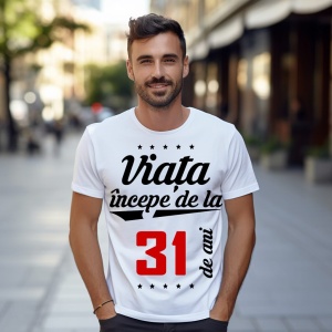 Bărbat zâmbitor purtând un tricou alb cu mesajul 'Viața începe de la 31 de ani', stând pe o stradă urbană însorită.