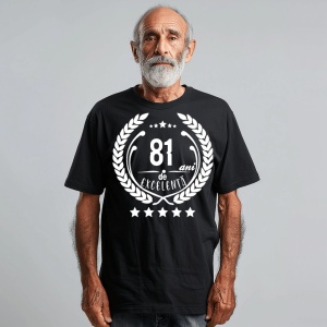 Un bărbat zâmbitor purtând un tricou negru cu textul ''81 de ani de excelență'', înconjurat de o coroană de lauri.