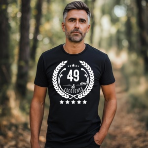 Un bărbat zâmbitor purtând un tricou negru cu textul ''49 de ani de excelență'', înconjurat de o coroană de lauri.