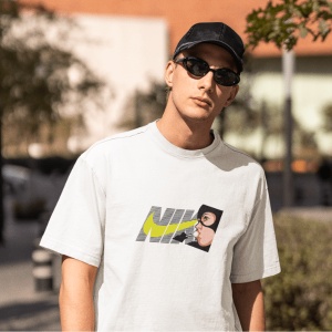 Tânăr cu ochelari de soare și șapcă neagră purtând un tricou alb cu un design Nike stilizat, incluzând un personaj animat lângă logo(Catwoman). Fundal urban ușor neclar, cu elemente de vegetație și clădiri.