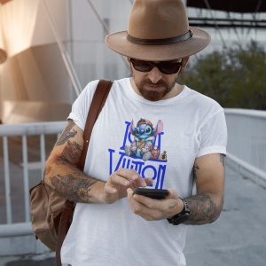 Bărbat cu pălărie și ochelari de soare purtând un tricou alb cu un personaj animat albastru(Stitch) stând pe un logo stilizat de Louis Vuitton, verificând telefonul, cu un rucsac maro pe umăr și tatuaje pe braț.