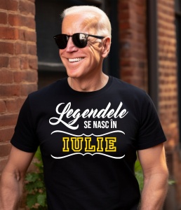 Un bărbat cu păr alb, purtând ochelari de soare și un tricou negru cu mesajul: „Legendele se nasc în Iulie”. Bărbatul zâmbește în timp ce stă lângă un zid de cărămidă, într-un cadru urban.