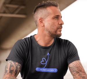 Bărbat cu tatuaje pe brațe purtând un tricou negru cu un design minimalist al unei săgeți în formă de inimă și textul 'CEL MAI BUN TATA' într-o bară de căutare stilizată. Fundalul este neutru, cu o structură modernă deasupra.