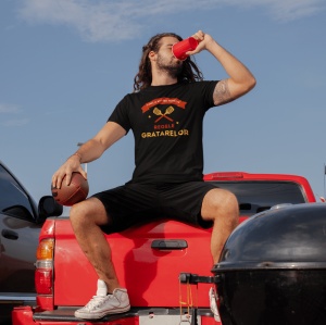 Bărbat relaxat stând pe marginea unui camion roșu, purtând un tricou negru cu mesajul 'Familia mă numește Regele Grătarelor,' decorat cu grafică de ustensile de grătar. Într-o mână ține o minge de fotbal american, iar în cealaltă o băutură în pahar roșu, 