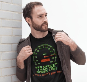 Un bărbat poartă un tricou negru cu un design ce afișează un vitezometru și mesajul amuzant 'Yes officer, I saw the speed limit, I just didn't see you' în text verde și roșu. Bărbatul poartă o jachetă și stă lângă un perete de cărămidă deschisă la culoare