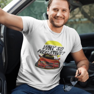 Bărbat zâmbitor purtând un tricou alb cu un design grafic ce prezintă o mașină sport roșie și textul 'Why so negative?' pe fundal galben, stând pe scaunul unei mașini și ținând o pereche de ochelari de soare în mână.
