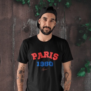 Bărbat cu barbă purtând o șapcă neagră pe spate și un tricou negru cu textul 'PARIS 1980' în roșu și albastru, pe fundal de lemn decorat cu frunze verzi.