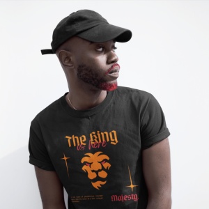 Un bărbat poartă un tricou negru cu inscripția 'The King is here' și o grafică a unui leu stilizat în portocaliu. El are o șapcă neagră și barba parțial colorată în roșu, privind lateral. Fundalul este simplu, alb.