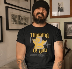 Un bărbat cu barbă și căciulă neagră zâmbește în timp ce poartă un tricou negru cu un design vintage. Designul conține un înger stilizat și textul 'Thinking of You' scris într-un font gotic galben. Sub imaginea îngerului apare un mesaj mic de afecțiune. B