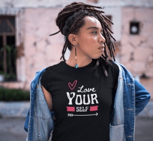 Fată cu părul dreadlocks purtând un tricou negru cu textul 'Love Yourself' și un desen cu inimă și săgeată, îmbrăcată într-o jachetă de blugi pe umeri, într-un cadru urban.