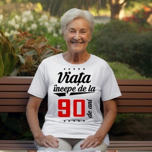 O femeie purtând un tricou alb cu textul  'Viața începe de la 90 de ani'. Fundal din lemn cu diferite nuanțe de maro și verde.