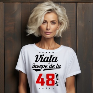 O femeie purtând un tricou alb cu textul  'Viața începe de la 48 de ani'. Fundal din lemn cu diferite nuanțe de maro și verde.