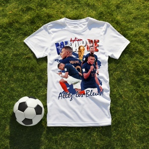 Tricou alb cu un design dedicat lui Kylian Mbappé, incluzând imagini ale fotbalistului, textul 'Allez les Bleus!' și steagul Franței, așezat pe iarbă alături de o minge de fotbal.