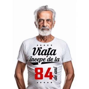 Bărbat în vârstă cu barbă albă, purtând un tricou alb cu mesajul 'Viața începe de la 84 de ani', pe un fundal alb simplu.