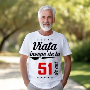 Bărbat în vârstă cu barbă albă, zâmbitor, purtând un tricou alb cu mesajul 'Viața începe de la 51 de ani', stând într-un parc luminat de soare.