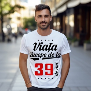 Bărbat zâmbitor purtând un tricou alb cu mesajul 'Viața începe de la 39 de ani', stând pe o stradă urbană însorită.