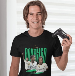 Un tânăr zâmbitor, ținând un controller de joc video, poartă un tricou negru cu design grafic dedicat jucătorului de fotbal Rodrygo, cu imagini ale acestuia și numele său scris cu litere verzi.