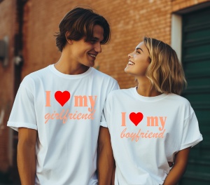 Un bărbat și o femeie purtând tricouri albe cu mesaje romantice. Tricoul bărbatului scrie 'I ❤️ my girlfriend', iar tricoul femeii scrie 'I ❤️ my boyfriend', privindu-se zâmbitori pe un fundal de clădire cu cărămidă.