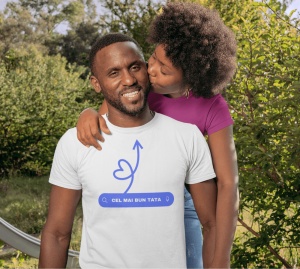 Bărbat zâmbitor purtând un tricou alb cu design minimalist al unei săgeți în formă de inimă și textul 'CEL MAI BUN TATA' într-o bară de căutare stilizată, în timp ce o persoană dragă îl îmbrățișează și îl sărută pe obraz. Fundalul este un peisaj natural, 