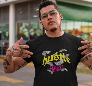 Bărbat cu tatuaje pe mâini și față, purtând un tricou negru cu design care include textul 'hustle HARD' și imagini cu bancnote. Poartă ochelari și arată cu degetele spre tricou, pe un fundal urban în fața unei clădiri cu geamuri mari.
