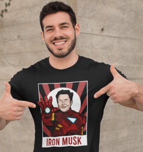 Bărbat zâmbitor și cu barbă, purtând un tricou negru cu un design amuzant care îl arată pe Elon Musk în costumul lui Iron Man, cu textul 'IRON MUSK' dedesubt. Bărbatul indică cu degetele spre designul tricoului, pe un fundal de perete texturat.