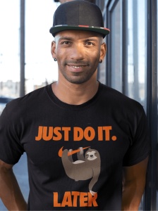 Un bărbat zâmbitor poartă un tricou negru cu un mesaj amuzant în portocaliu care spune 'JUST DO IT. LATER' și o ilustrație a unui leneș agățat de o ramură.