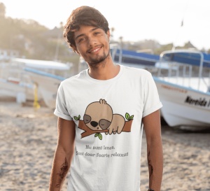 Un tânăr poartă un tricou alb cu o ilustrație drăguță a unui leneș care doarme pe o creangă, alături de textul 'Nu sunt leneș, Sunt doar foarte relaxat'. Tânărul zâmbește, iar fundalul arată o plajă cu bărci ancorate.