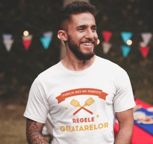 Bărbat zâmbitor purtând un tricou alb cu mesajul 'Familia mea mă numește Regele Grătarelor,' decorat cu grafică de ustensile de grătar. În fundal, sunt vizibile decorațiuni colorate de petrecere, creând o atmosferă festivă în aer liber.