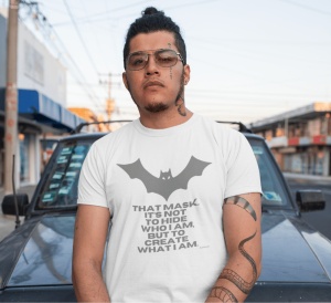 Bărbat cu ochelari de vedere și tatuaje pe gât și braț, purtând un tricou alb cu un design inspirat de Batman, reprezentând o siluetă de liliac și textul: 'THAT MASK. IT'S NOT TO HIDE WHO I AM, BUT TO CREATE WHAT I AM.' În spate se află o mașină și un pei