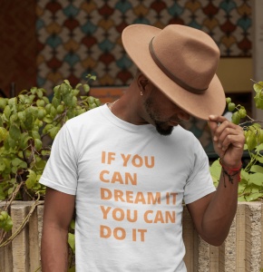 Bărbat purtând un tricou alb cu mesajul motivațional 'IF YOU CAN DREAM IT YOU CAN DO IT' scris cu litere portocalii și o pălărie maro, înclinând capul în jos. Fundalul include o plantă și un perete decorativ cu modele geometrice colorate.